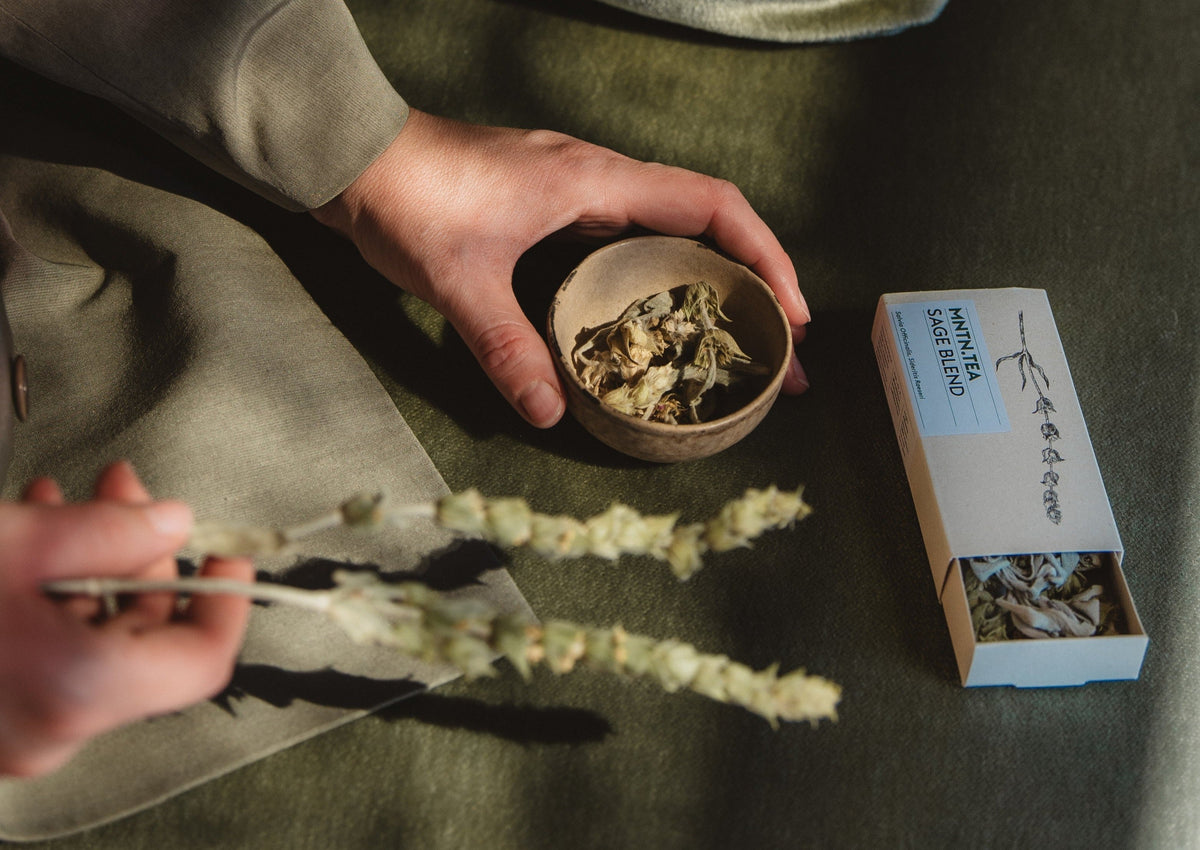 Handen met een kommetje gedroogde bergthee en salie, naast het doosje Sage Blend